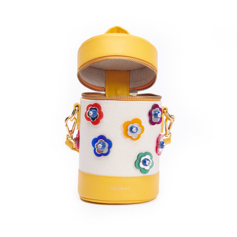 Bolsa cilíndrica em tecido bege com detalhes em couro amarelo. Alça longa removível e aplicações frontais de flores coloridas em relevo com miolo em cristal. Fechamento em zíper metálico dourado.