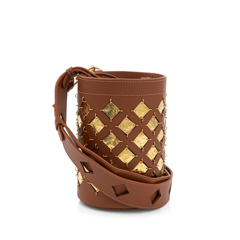 Bolsa bucket em couro marrom com aplicações geométricas quadradas em metal dourado. Possui alça tiracolo larga com recortes geométricos e fechamento superior com amarração interna.