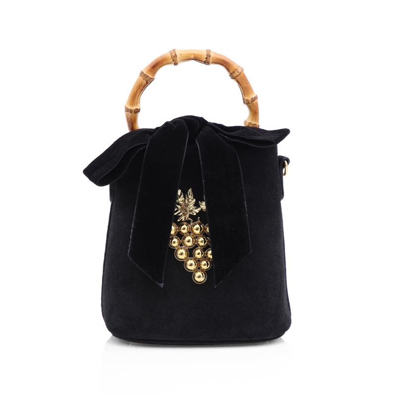 Bolsa bucket em veludo preto com alça de bambu, laço de veludo no topo e detalhe frontal de cacho de uvas dourado com folhas metálicas.