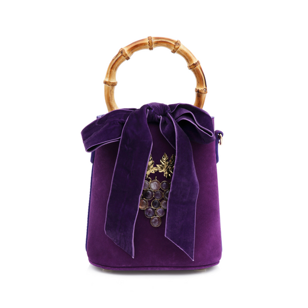 Bolsa bucket em veludo roxo com alça de bambu, laço de veludo no topo e detalhe frontal de cacho de uvas dourado com pedras escuras e folhas metálicas.