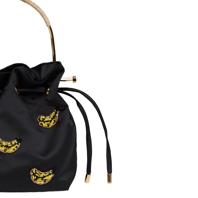 Bolsa bucket luxuosa em seda preta, decorada com bananas em cristais, alça estruturada dourada com detalhe em rattan e fecho em cordão.
