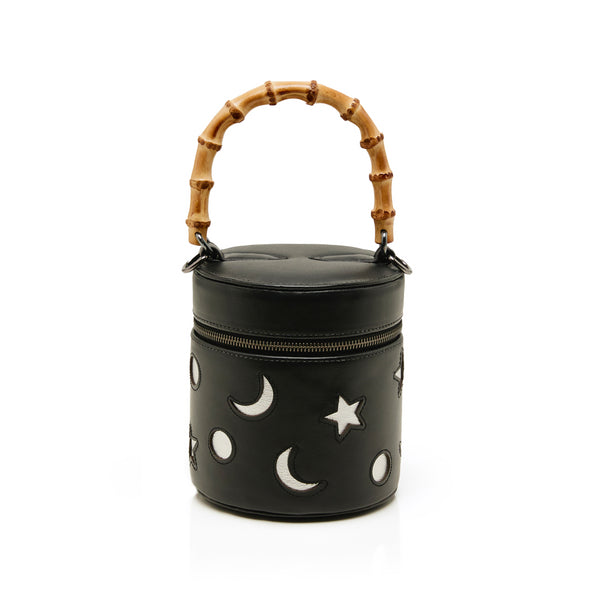 Bolsa cilíndrica preta em couro com bordados de luas e estrelas, fechamento em zíper e alça estruturada em bambu.