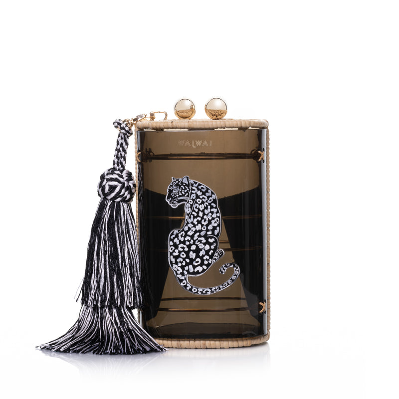 Bolsa clutch estruturada em acrílico preto translúcido, acabamento em rattan natural, estampa de onça branca central e franja longa em seda preta e branca, com fecho em esferas douradas.