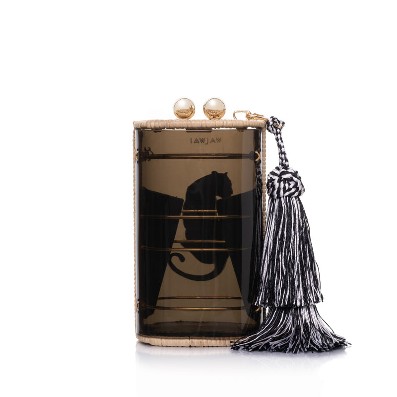 Bolsa clutch estruturada em acrílico preto translúcido, acabamento em rattan natural, estampa de onça branca central e franja longa em seda preta e branca, com fecho em esferas douradas.
