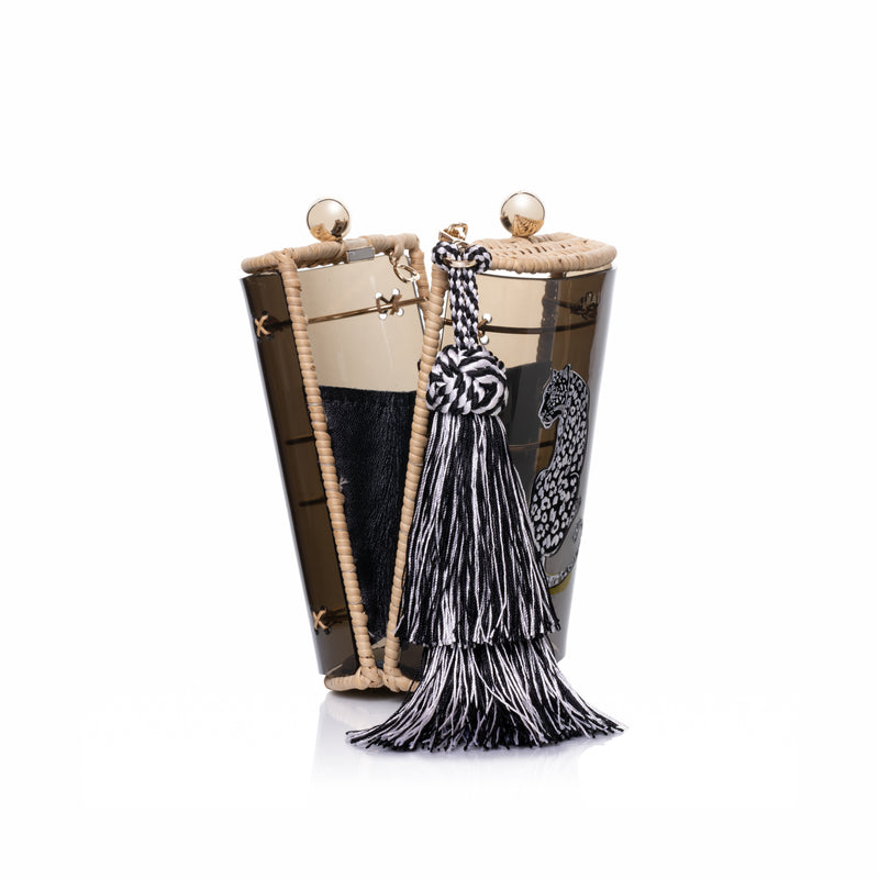 Bolsa clutch estruturada em acrílico preto translúcido, acabamento em rattan natural, estampa de onça branca central e franja longa em seda preta e branca, com fecho em esferas douradas.