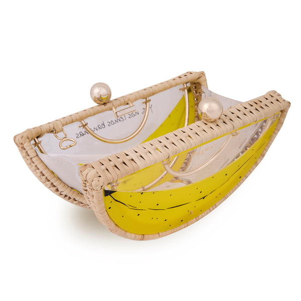 Clutch em formato meia-lua com estrutura em rattan e acrílico transparente, estampa frontal de banana amarela estilizada e fecho metálico dourado no topo.