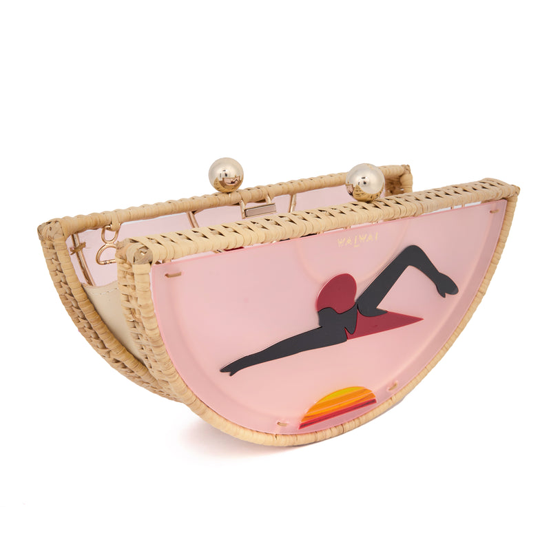 Clutch em formato meia-lua com estrutura em rattan e acrílico rosa translúcido, estampa estilizada de mulher deitada na praia com sol poente ao fundo, e fecho metálico dourado no topo.