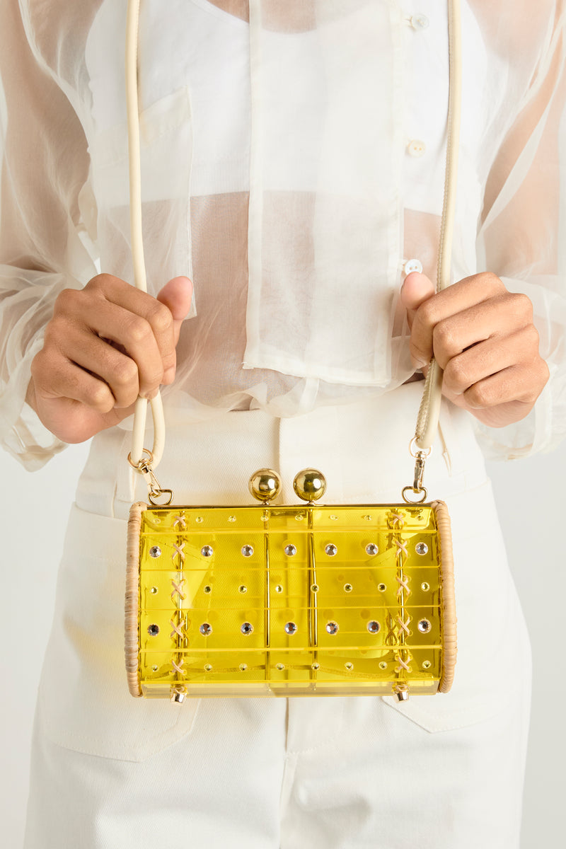 Clutch estruturada em rattan com corpo em acrílico amarelo translúcido, aplicação de cristais circulares e fecho metálico dourado no topo.