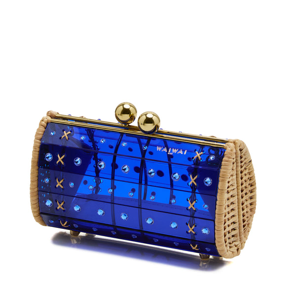 Clutch em rattan com corpo em acrílico azul translúcido, cristais circulares aplicados e fecho metálico dourado no topo, fotografada de frente.