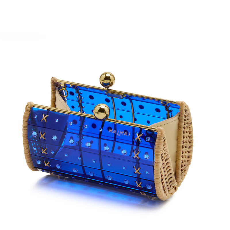 Clutch artesanal em rattan, corpo em acrílico azul translúcido com cristais redondos e fecho metálico dourado, ângulo frontal.