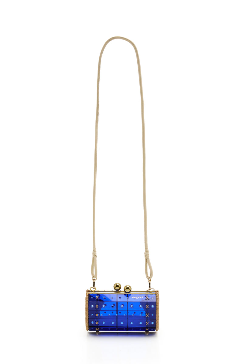 Clutch estruturada em rattan natural com corpo em acrílico azul translúcido, aplicação de cristais e fecho dourado superior, vista de frente.