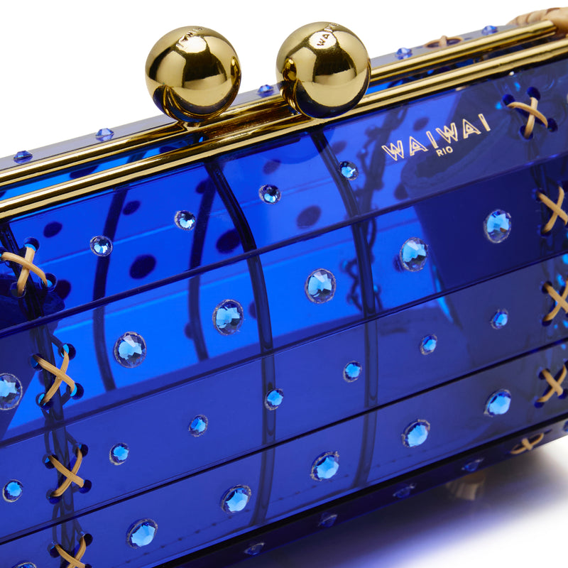 Clutch em rattan trançado com corpo em acrílico azul translúcido, cristais circulares aplicados e fecho metálico dourado, tomada frontal.
