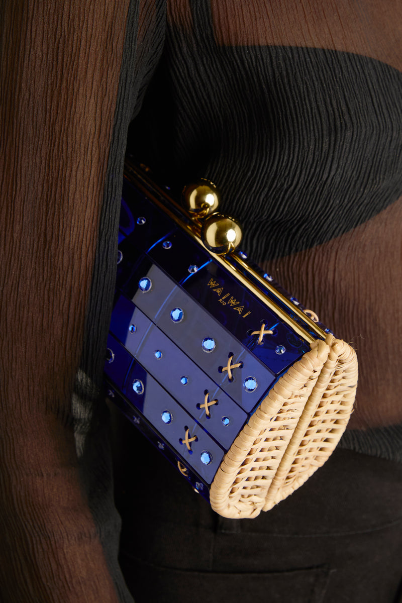 Clutch em rattan com corpo de acrílico azul translúcido, aplicação de cristais circulares e fecho metálico dourado no topo, imagem em ângulo frontal.