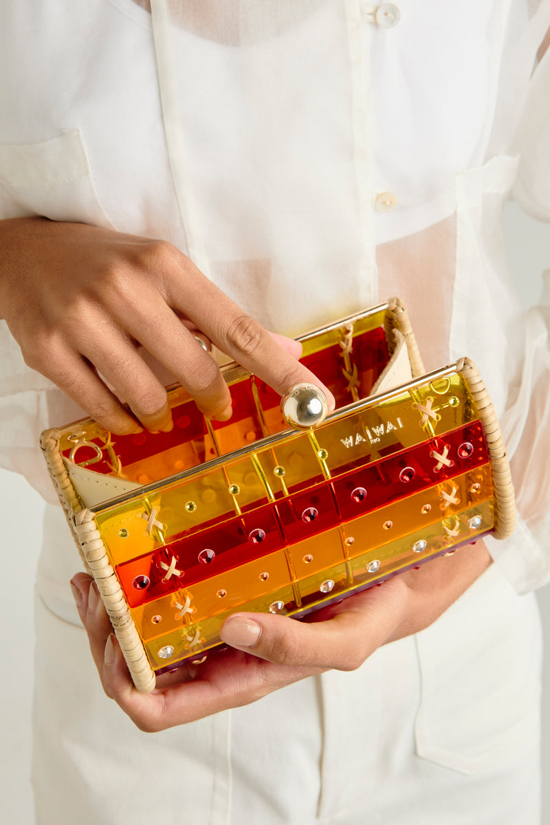 Clutch em rattan trançado com corpo de acrílico translúcido listrado em vermelho e amarelo, cristais aplicados e fecho metálico dourado.