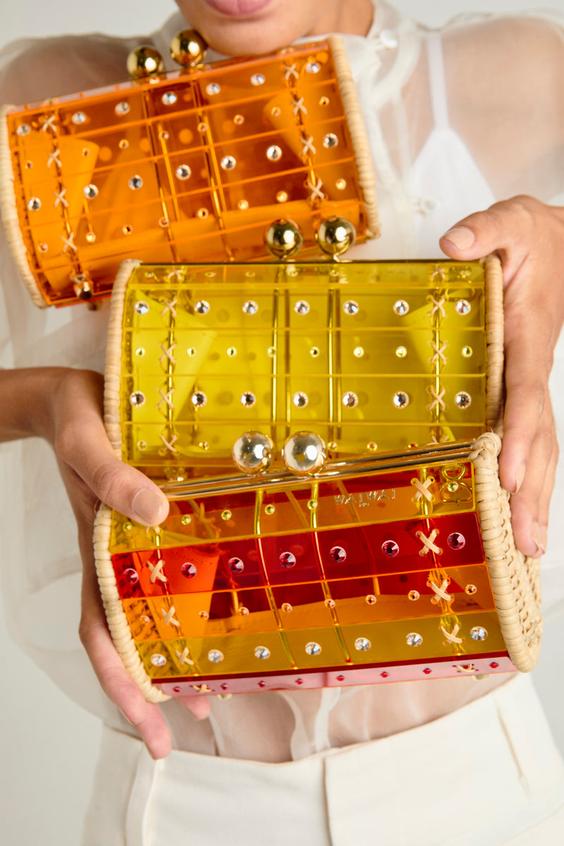 Clutch estruturada em rattan, corpo em acrílico translúcido listrado bicolor vermelho e amarelo, aplicações de cristais e fecho dourado metálico.