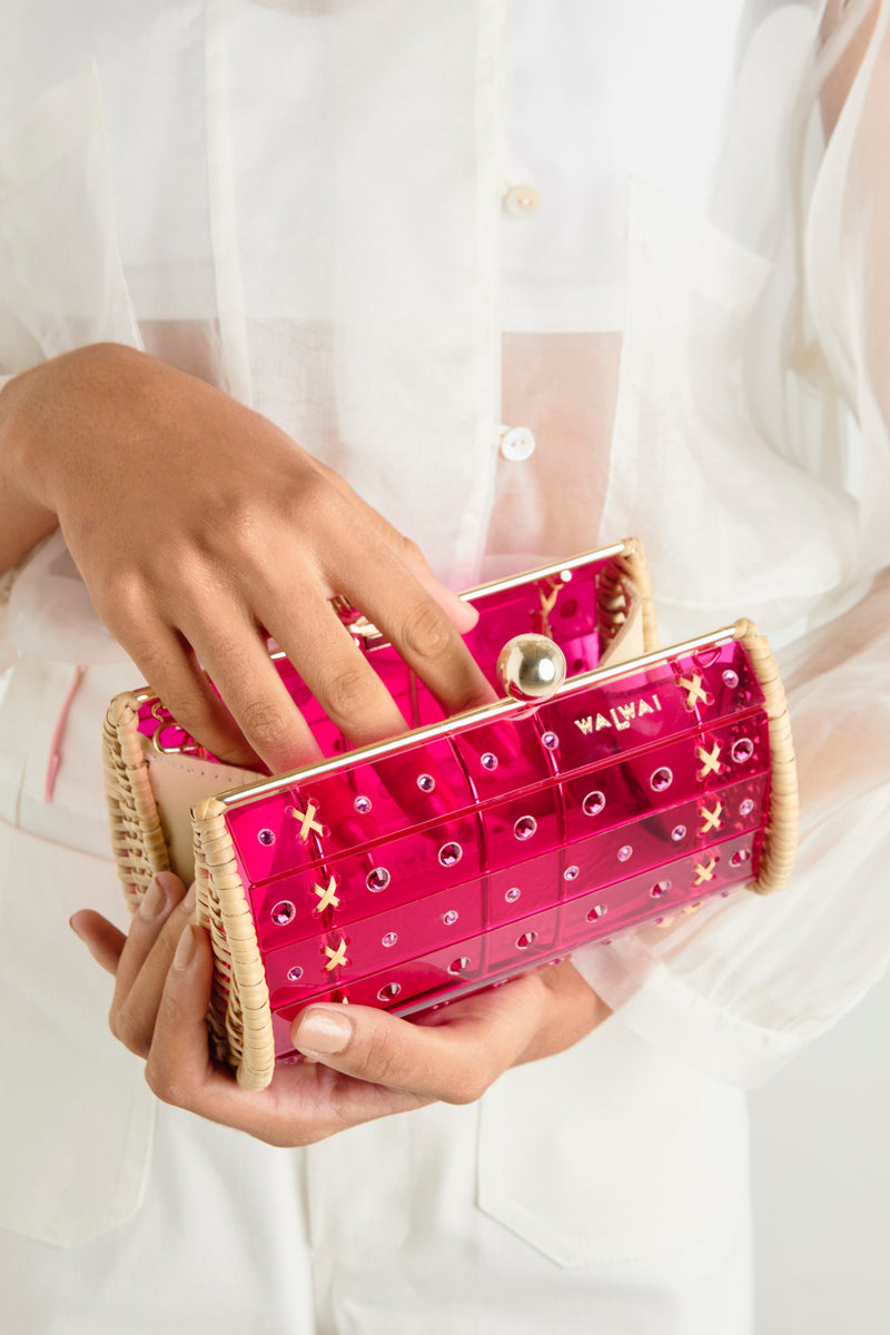 Clutch em rattan trançado com corpo em acrílico rosa translúcido, decorada com cristais circulares e fecho dourado no topo.