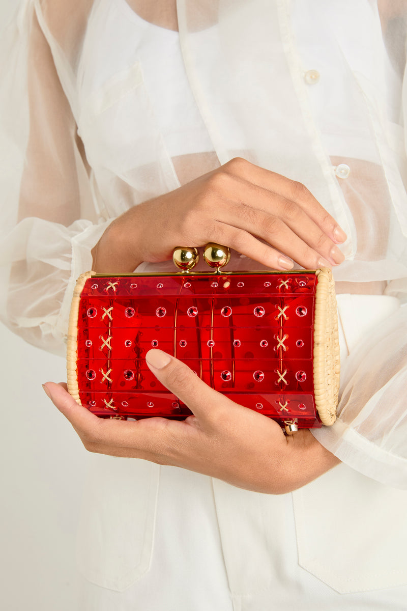 Clutch estruturada em rattan com corpo em acrílico vermelho translúcido, aplicação de cristais circulares e fecho metálico dourado no topo.