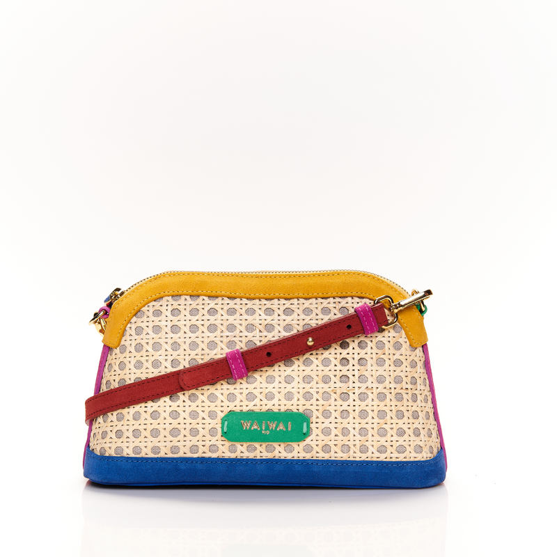 Bolsa estruturada com corpo em palha tramada natural, acabamentos em camurça colorida — amarelo, azul e pink — e alça tiracolo regulável em couro vermelho. Detalhe de plaquinha verde com logo na frente.