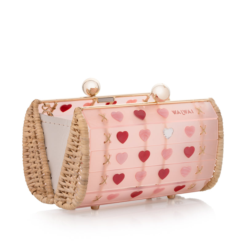 Bolsa de acrílico rosa translúcido com estrutura em metal dourado revestida de vime e estampa de corações vermelhos e rosas.