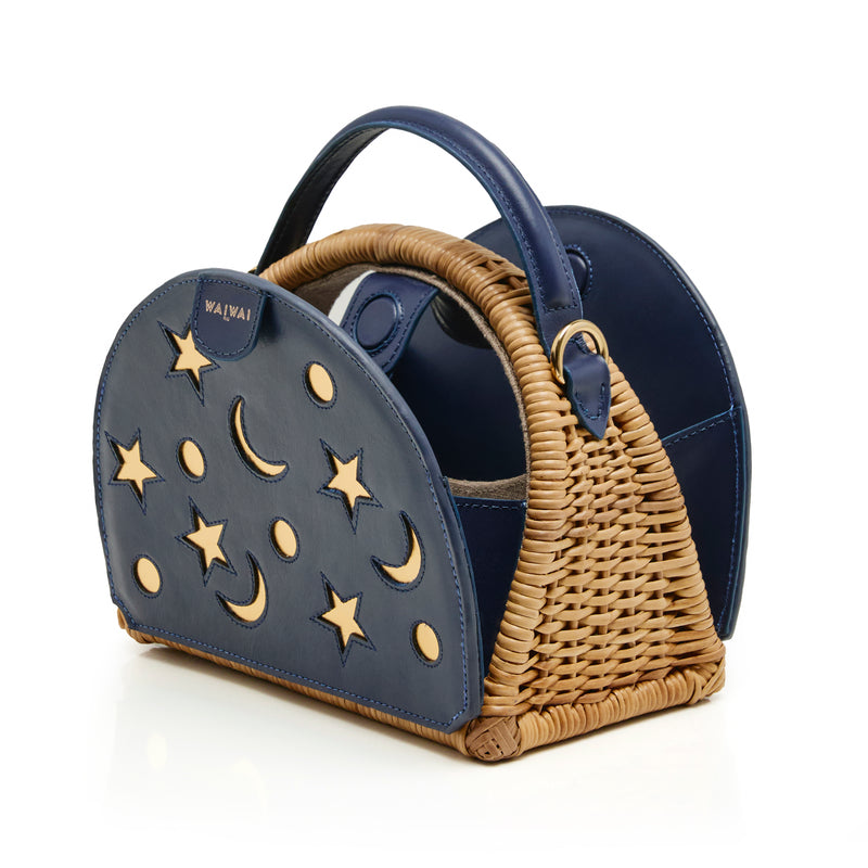 Bolsa estruturada em rattan com laterais em couro azul marinho, recortes de luas e estrelas, e alça superior em couro.