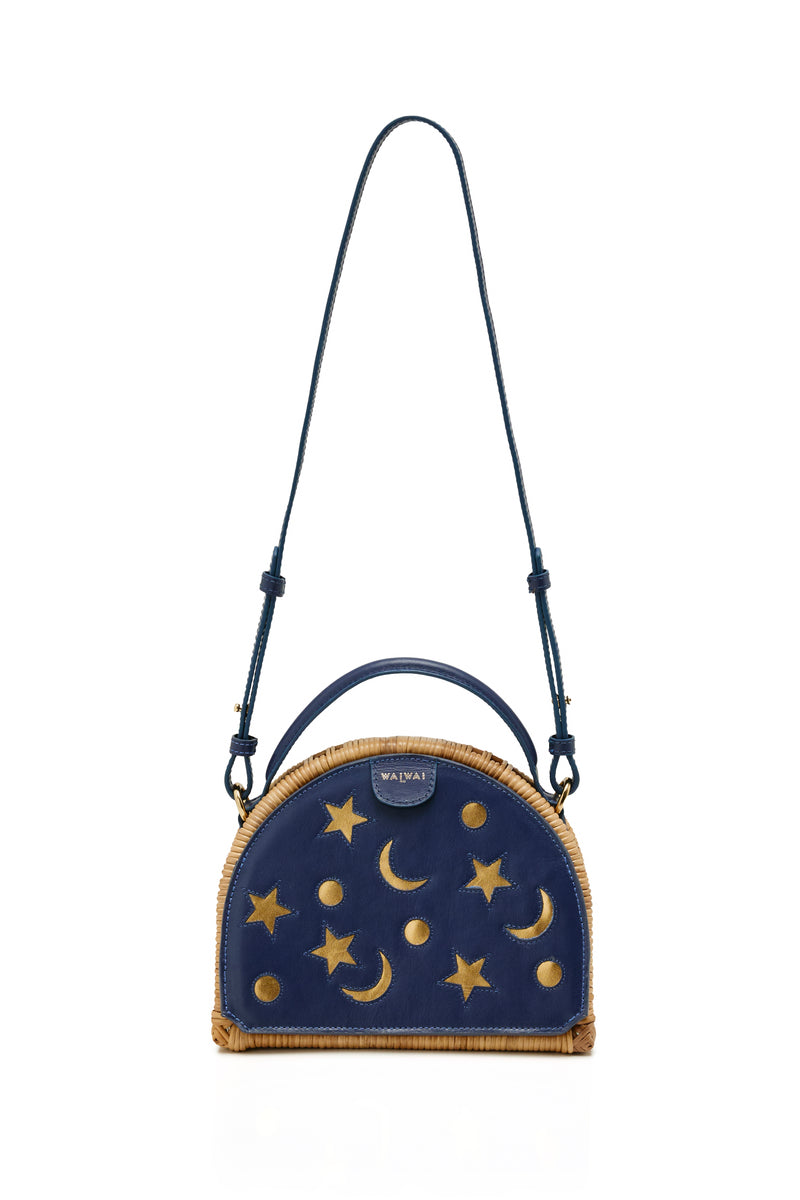 Bolsa estruturada em rattan com laterais em couro azul marinho, recortes de luas e estrelas, e alça superior em couro.