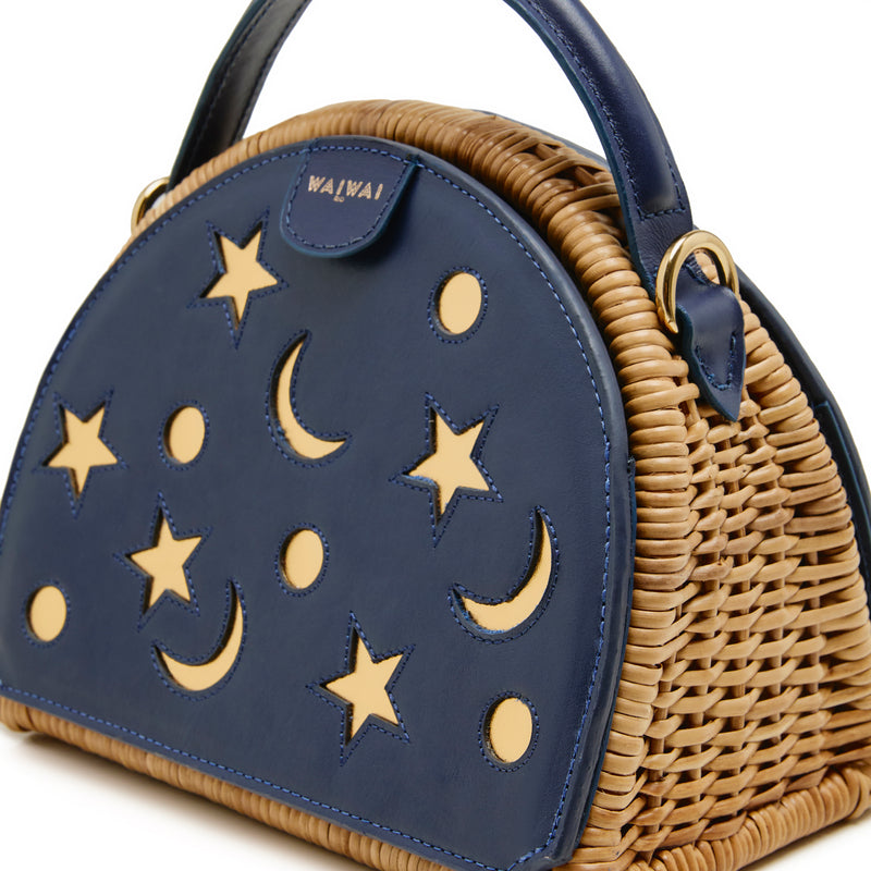 Bolsa estruturada em rattan com laterais em couro azul marinho, recortes de luas e estrelas, e alça superior em couro.