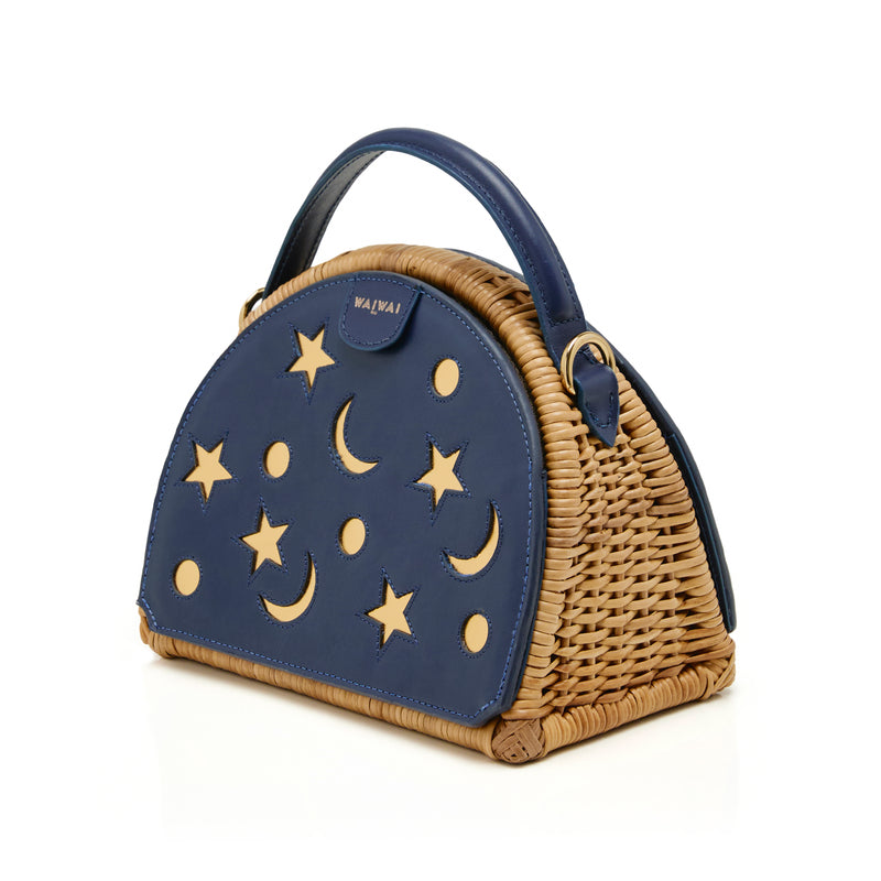 Bolsa estruturada em rattan com laterais em couro azul marinho, recortes de luas e estrelas, e alça superior em couro.