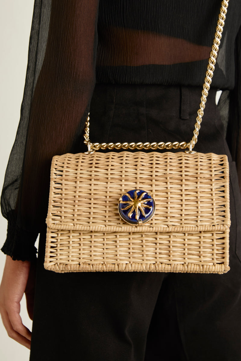 Bolsa grande em rattan natural estruturado, com detalhe azul marinho e sol dourado central, alça longa de corrente metálica dourada.