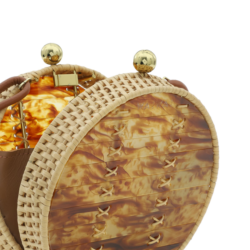 Bolsa redonda em acrílico marmorizado caramelo, com alça longa em couro marrom, detalhes em palha trançada nas bordas e fecho esférico dourado.