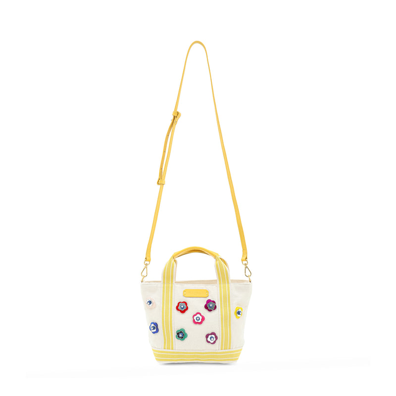 Bolsa em tecido bege estruturado com alças de mão e tiracolo em amarelo, decorada com aplicações de flores multicoloridas em relevo. Detalhe de etiqueta frontal em couro amarelo com logo.