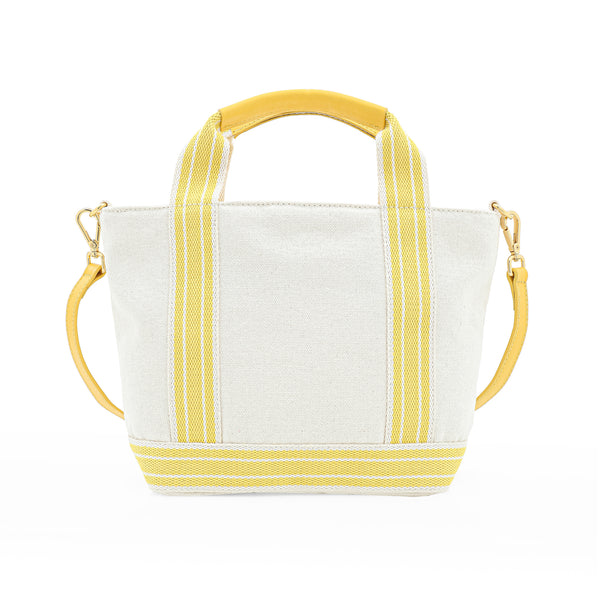 Bolsa estruturada em tecido bege com alças de mão e tiracolo em amarelo, aplicações florais multicoloridas em relevo e detalhe frontal em couro amarelo com logo.