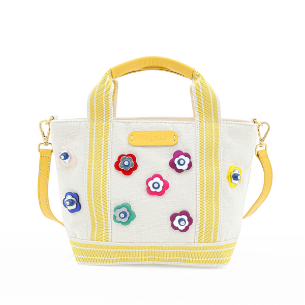 Bolsa em tecido bege estruturado com alças de mão e tiracolo em amarelo, decorada com aplicações de flores multicoloridas em relevo. Detalhe de etiqueta frontal em couro amarelo com logo.