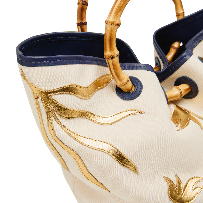 Bolsa tote em tecido cru com bordados dourados, base em azul marinho e alça estruturada em bambu.