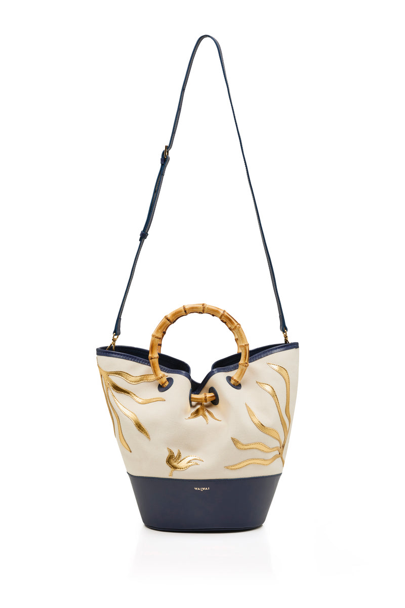 Bolsa tote em tecido cru com bordados dourados, base em azul marinho e alça estruturada em bambu.