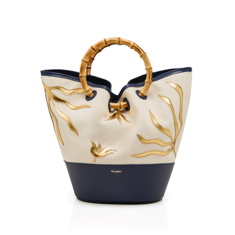 Bolsa tote em tecido cru com bordados dourados, base em azul marinho e alça estruturada em bambu.