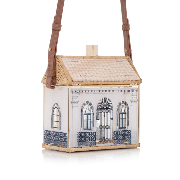 Bolsa autoral em acrílico branco e vime natural, com fachada colonial azul e estrutura dourada.