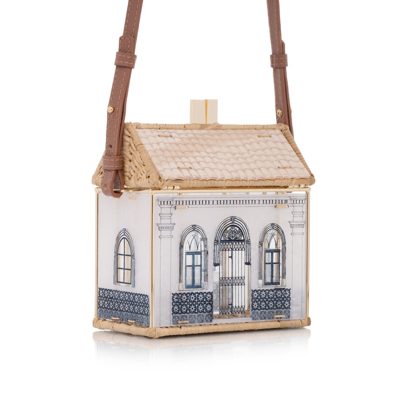 Bolsa autoral em acrílico branco e vime natural, com fachada colonial azul e estrutura dourada.