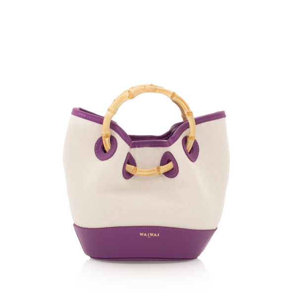 Bolsa bucket em canvas cru com detalhes em couro roxo. Possui alça de mão em bambu curvado e acabamento em couro na base e nas bordas superiores.
