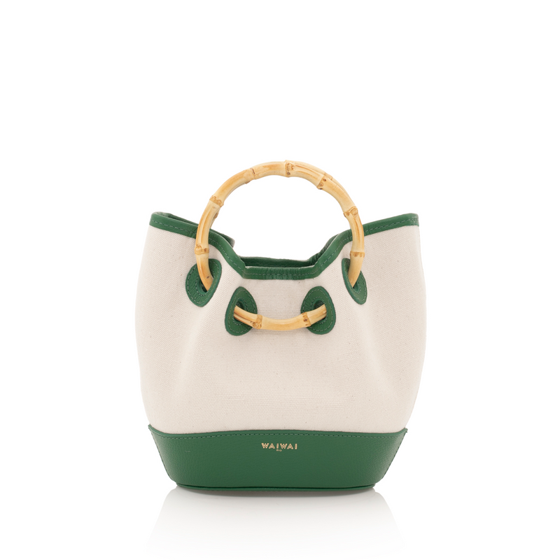 Bolsa bucket em canvas bege com acabamento em couro verde e alça de bambu natural. Elegância tropical e atemporal.
