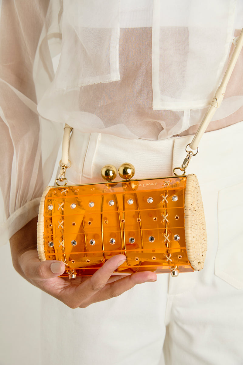 Clutch em acrílico laranja translúcido com design geométrico, acabamentos em palha nas laterais e fecho dourado no topo.
