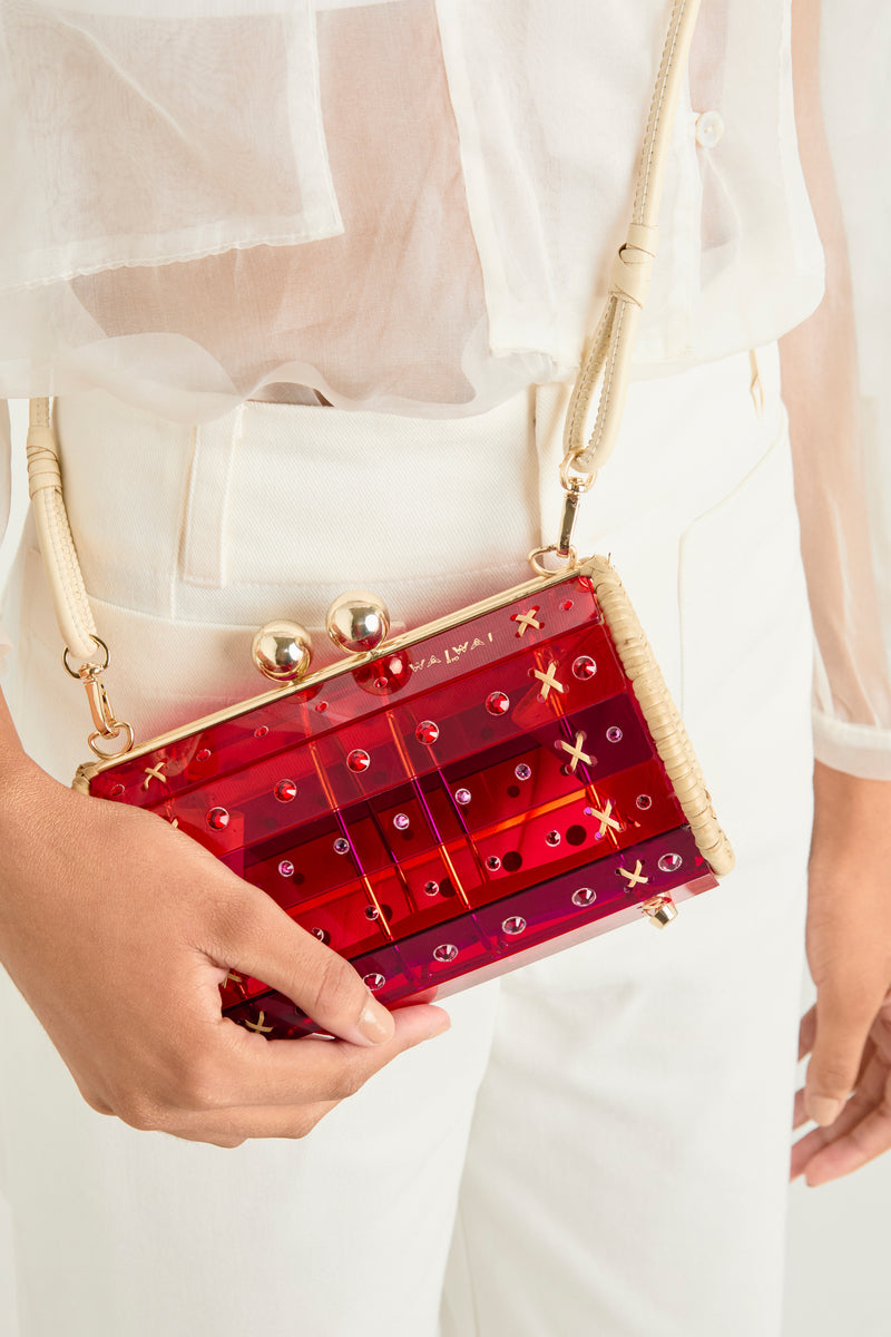 Clutch em acrílico vermelho translúcido com design geométrico, acabamentos laterais em palha e fecho dourado no topo.