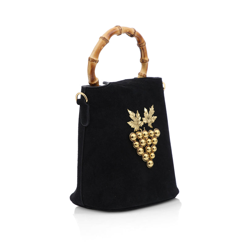 Bolsa bucket de veludo preto com alça em bambu, laço no topo em veludo e aplicação frontal de cacho de uvas dourado com folhas metálicas.