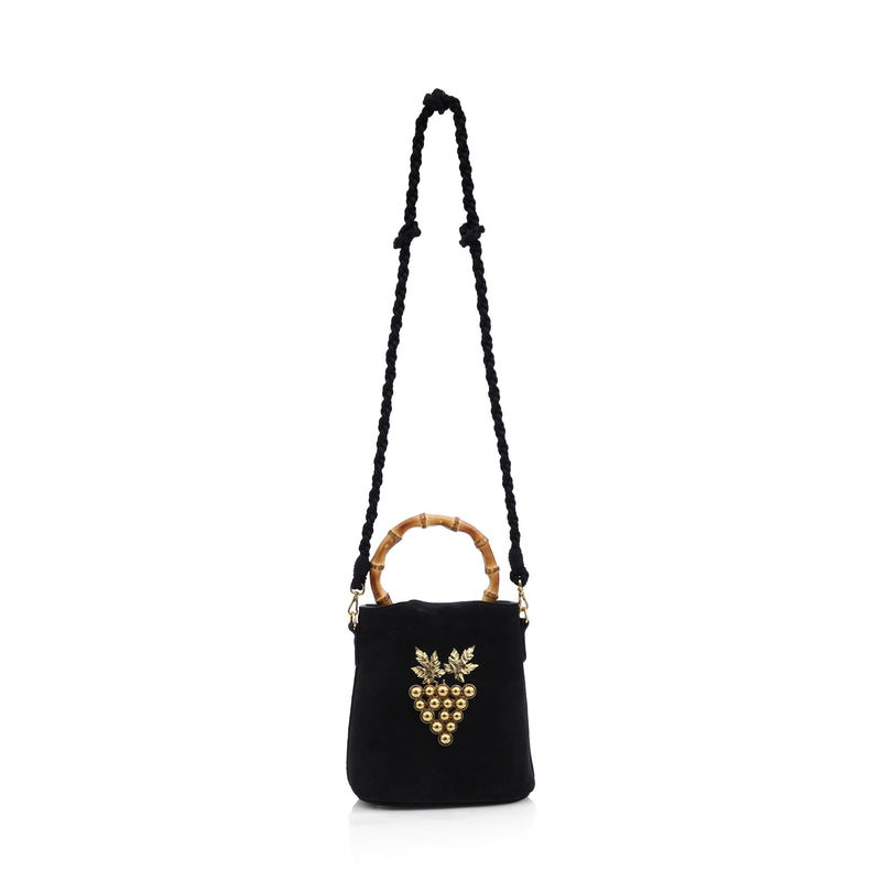 Bolsa bucket em veludo preto, alça rígida de bambu, laço no topo e detalhe frontal de cacho de uvas dourado com folhas metálicas.