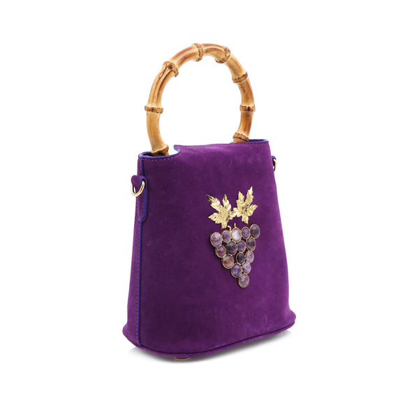Bolsa bucket de veludo roxo com alça em bambu, laço superior em veludo e aplicação frontal de cacho de uvas dourado com pedras escuras e folhas metálicas.