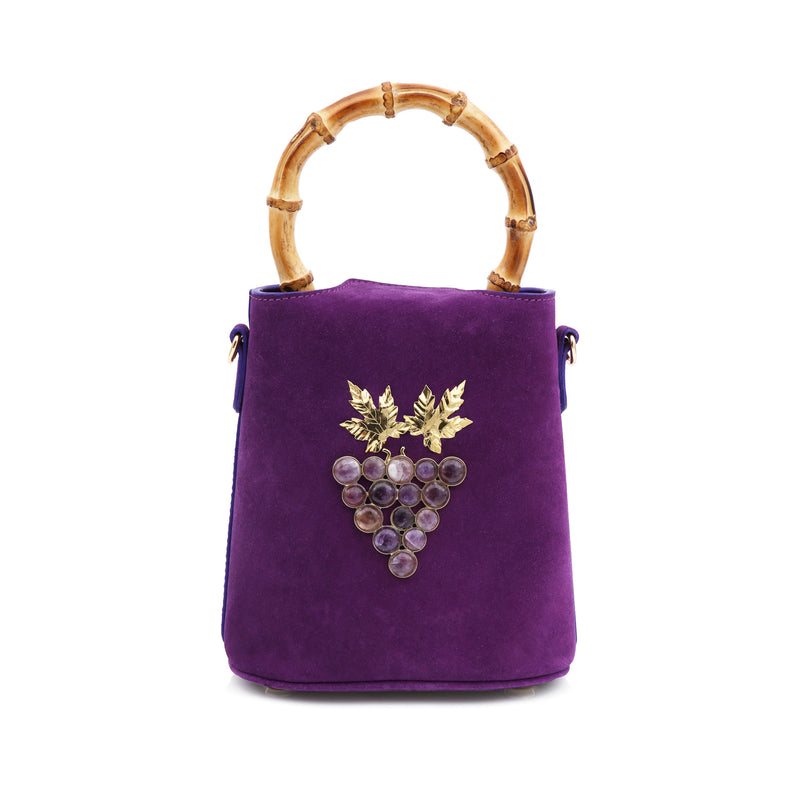 Bolsa bucket em veludo roxo, alça rígida de bambu, laço no topo e detalhe frontal de uvas douradas com pedras escuras e folhas metálicas.