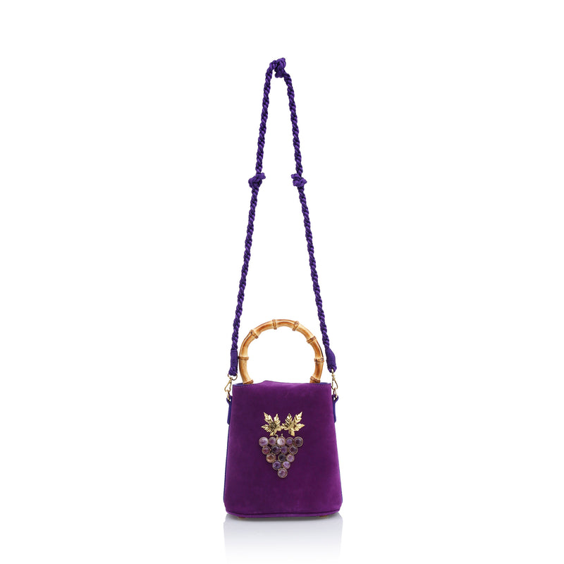 Bolsa bucket estruturada em veludo roxo com alça de bambu, laço de veludo superior e ornamento frontal de uvas douradas com pedras escuras e folhas metálicas.