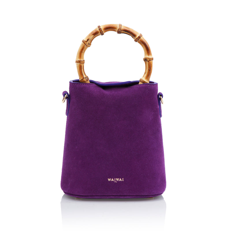 Bolsa bucket roxa em veludo, com alça em bambu, laço no topo e detalhe frontal de cacho de uvas dourado com pedras escuras e folhas metálicas.