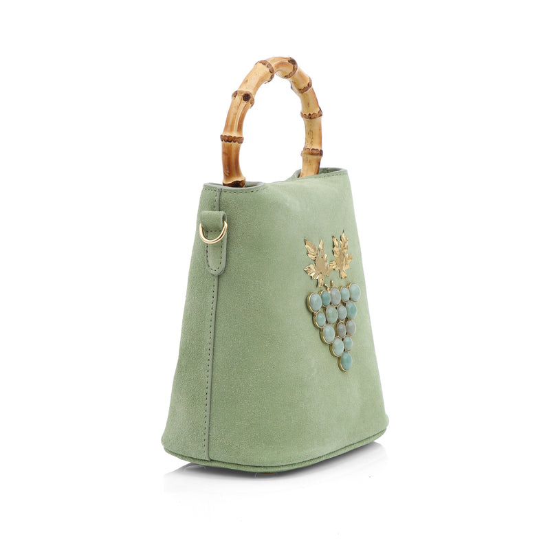 Bolsa bucket de camurça verde com alça de bambu natural e detalhe de cacho de uvas em pedras verdes com folhas metálicas douradas.