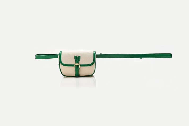Bolsa de canvas cru com acabamento em couro verde e fivela dourada, versátil para ser usada tanto como crossbody com alça longa quanto como pochete ajustada na cintura.