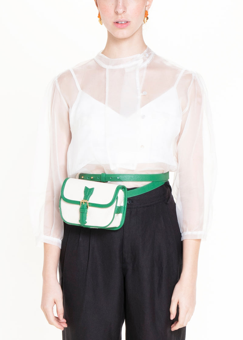 Bolsa de canvas cru com acabamento em couro verde e fivela dourada, versátil para ser usada tanto como crossbody com alça longa quanto como pochete ajustada na cintura.